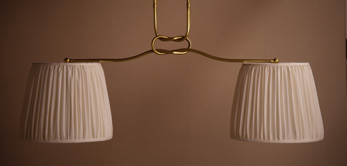 A modern two-light pendant chandelier with a polished brass-finished metal frame and two pleated white fabric drum shades, shown against a solid warm brown background.