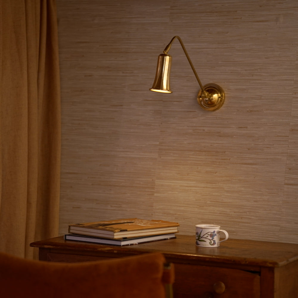 Belle Wall Sconce in brass on a textured wall with a wooden table and books below.