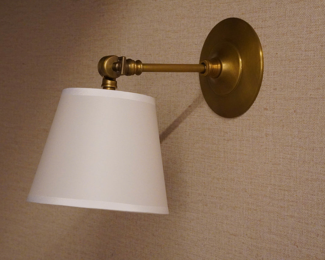A wall-mounted sconce with a white tapered lampshade, brass-colored metal arm and backplate, shown against a beige textured wall.