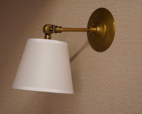 A wall-mounted sconce with a white tapered lampshade, brass-colored metal arm and backplate, shown against a beige textured wall.